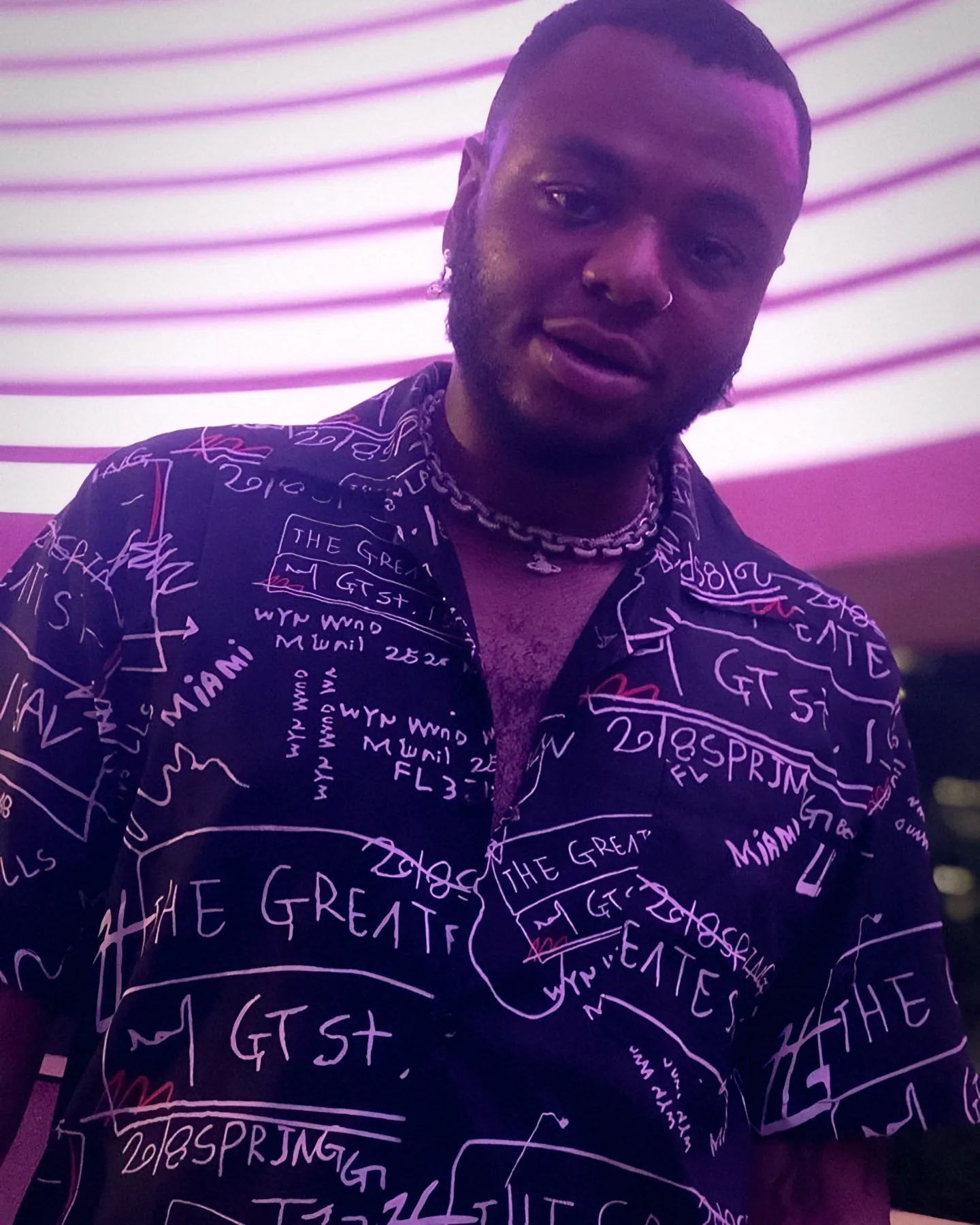Bobby Brown Jr. facing the camera in a black 'The Greatest' print shirt and chain under purple neon stripes